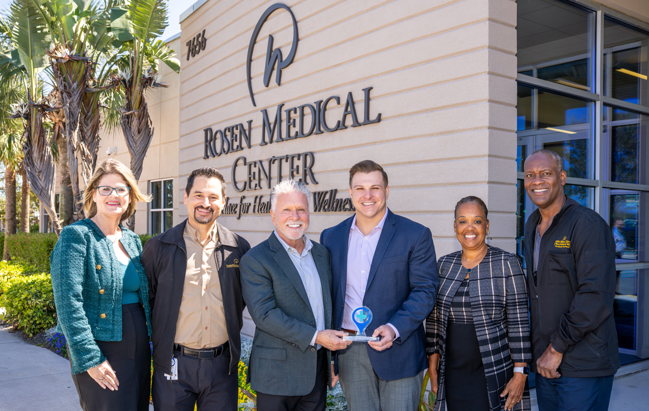 Four men and two women stand in front of the Rosen Medical Center, two gentlemen in the middle holding a small award. 