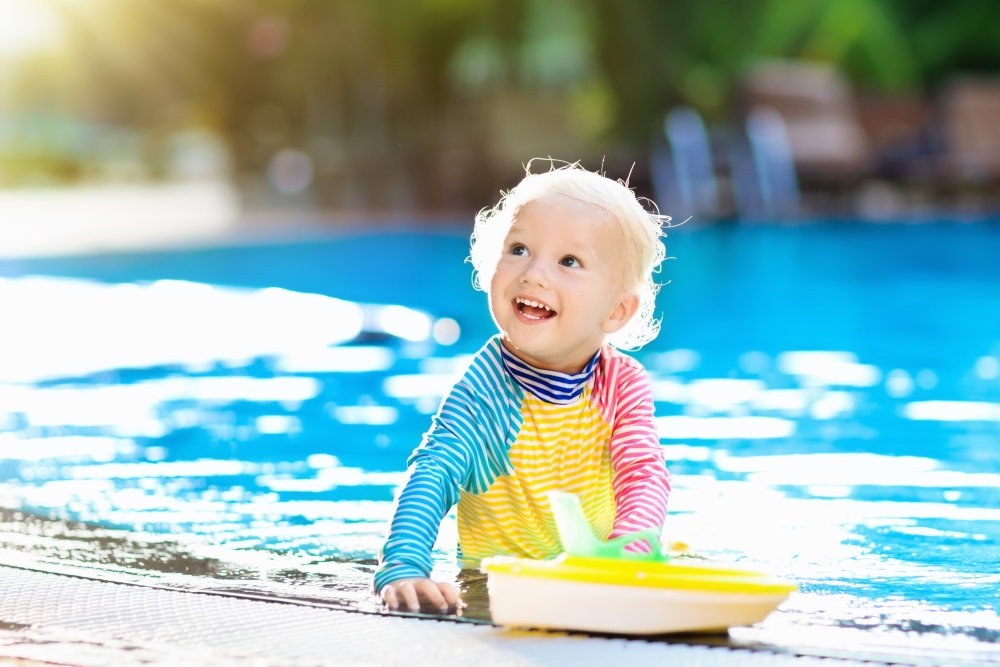 A child in long-sleeved protective swimwear smiles in a pool.