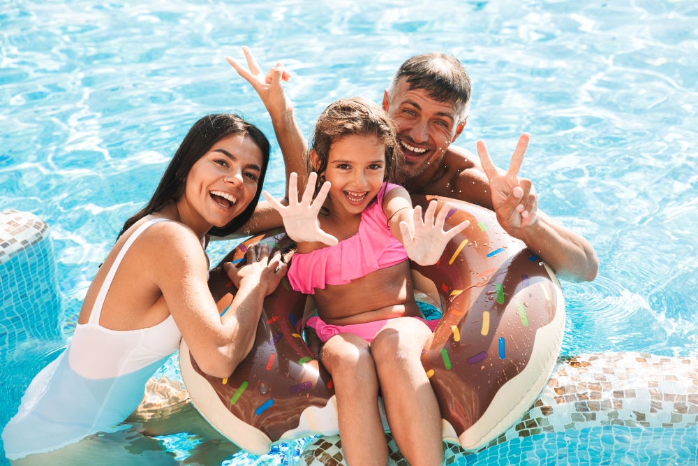 A mom, dad, and young girl smile and wave at the camera from a donut-shaped inner tube in a pool. 