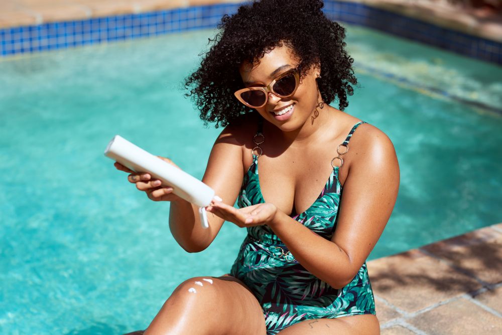 A woman in a bathing suit and sunglasses sitting beside a pool squeezes sunscreen into her hand. 