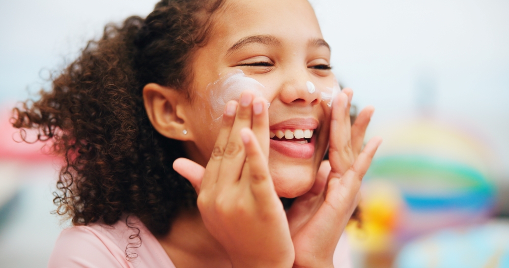 A young girl spreads sunscreen on her cheeks, smiling. 