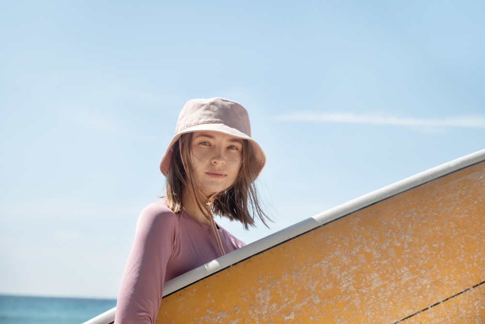 A woman wearing a hat carries a surfboard on the beach. 