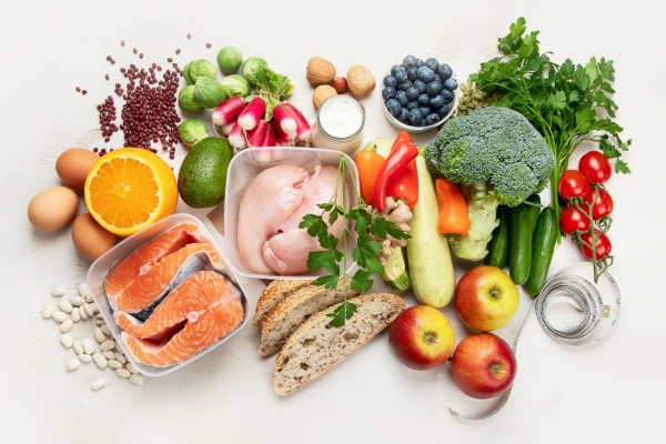 An array of healthy food against a white backdrop, including raw salmon, chicken breast, whole grain bread, fruits, and veggies.
