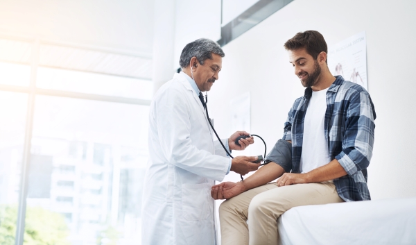 A man in a blue flannel shirt has his blood pressure taken by a male doctor in a white coat.