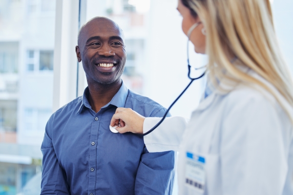 A man in a blue button down shirt smiles as a female doctor checks his heart rate with a stethoscope.