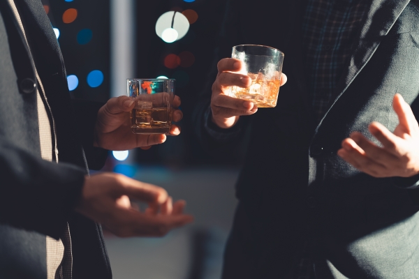 Close-up shot of two men in suits talking and holding whiskey glasses.