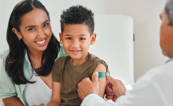 A boy sitting in a woman's lap smiles as a doctor places a green bandage on his arm where he received a vaccine.