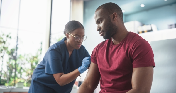 A man in a red t-shirt receives a vaccine in his arm from a nurse in blue scrubs and gloves.