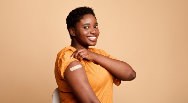 A woman in an orange shirt smiles and lifts her sleeve, revealing a band-aid on her arm. 