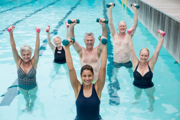 Five seniors in bathing suits stand behind an instructor, all of whom are standing waist-deep in a pool wearing bathing suits and with two dumbbells raised over their heads.