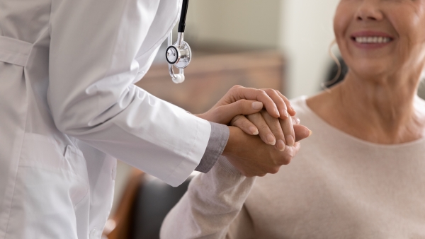 A close-up of a woman in a white lab coat holds the hand of an older woman.