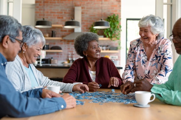 A group of five seniors smile and laugh seated around a table with puzzles pieces and white mugs.