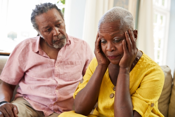 A man in a pink button down looks at a woman in a yellow shirt holding her temples with concern.