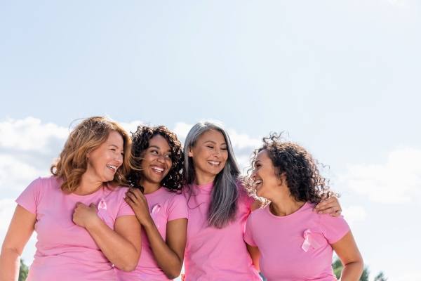 A diverse group of women in matching pink shirts and pink breast cancer ribbons embrace, smiling and laughing. 