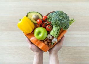 A heart-shaped bowl holding fruits, vegetables, salmon, and almonds.