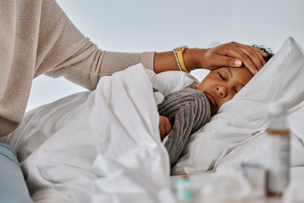 A woman rests her hand on the forehead of a child who lies sick in bed holding a stuffed toy.