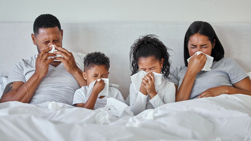 A mom, dad, and two children all sit under the covers of a bed, blowing their noses.