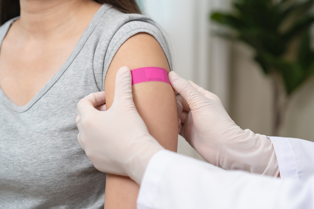 A close-up of a doctor's gloved hands placing a pink bandage on a woman's arm after getting a flu shot.