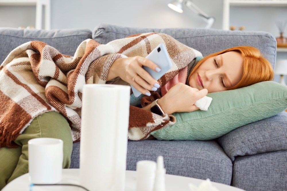 A red-headed woman lies on the couch, holding her phone and tissues.