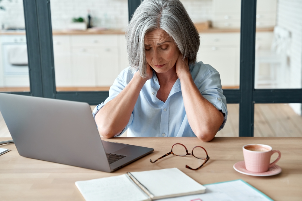 A woman sits at a desk in front of a laptop. rubbing her neck. 