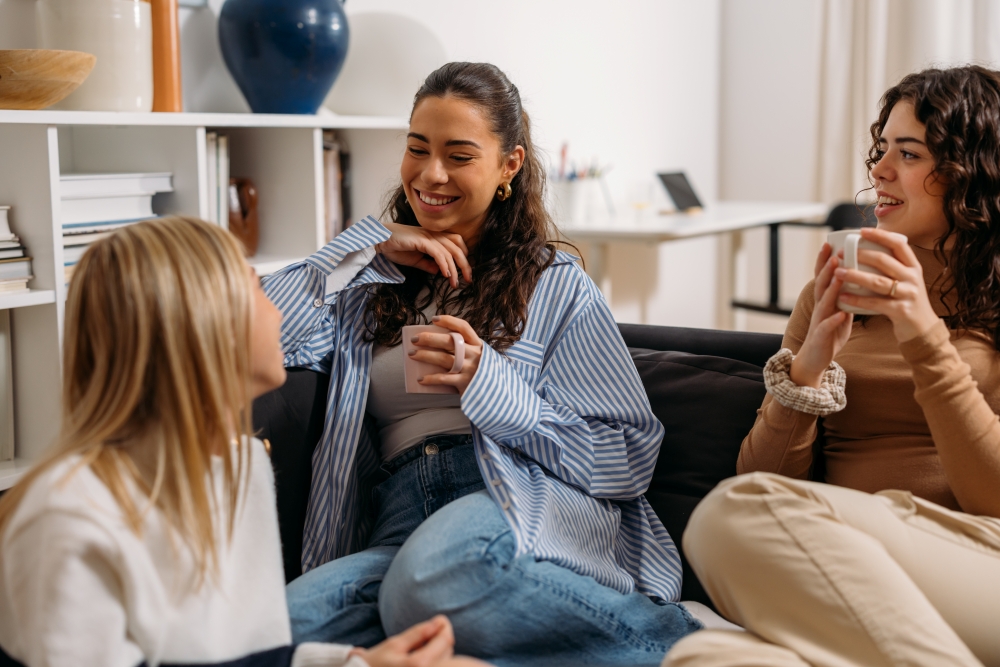 Three women smile, sitting on a sofa and holding cups of coffee. 