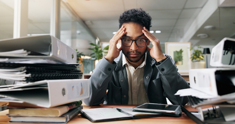A man in a jacket sits at a desk stacked with binders and books, his head resting in his hands. 