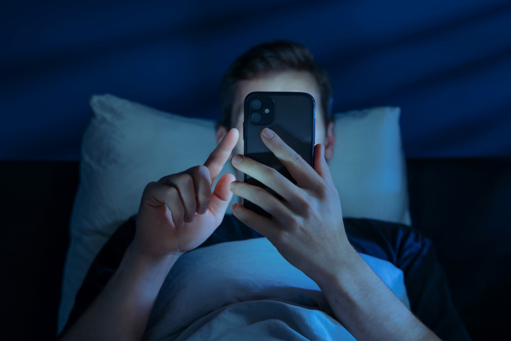 A man lies in bed in the dark, holding his phone in front of his face as he scrolls. 