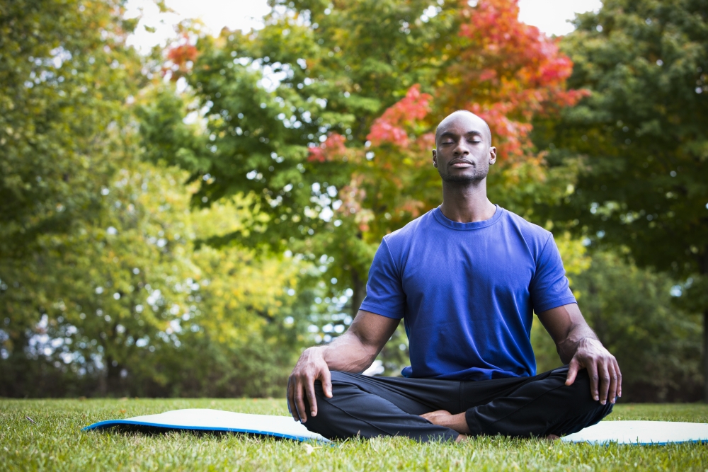 A physically fit man sits cross-legged on grass outdoors, his eyes closed and at peace.