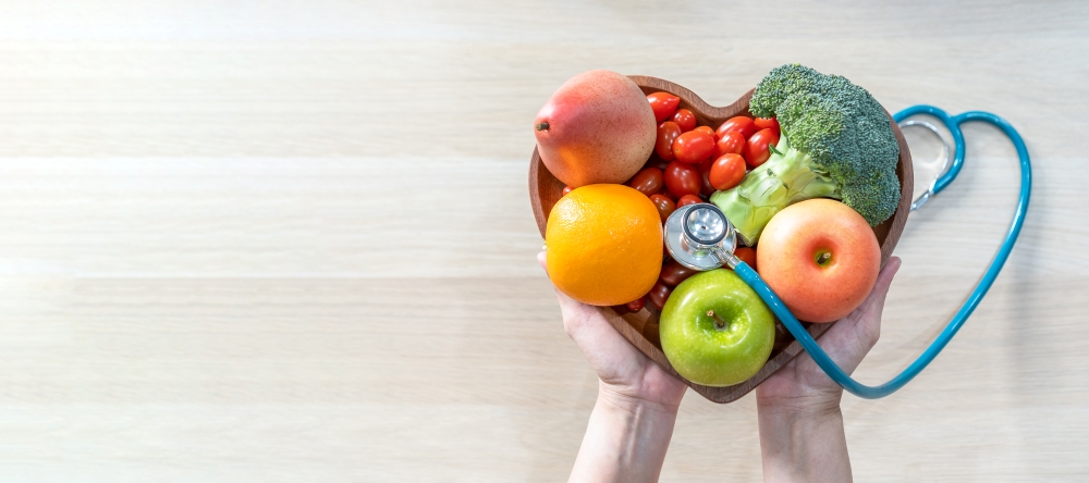 Hands hold a wooden bowl shaped like a heart filled with apples, broccoli, tomatoes, and a stethoscope also in the shape of a heart.