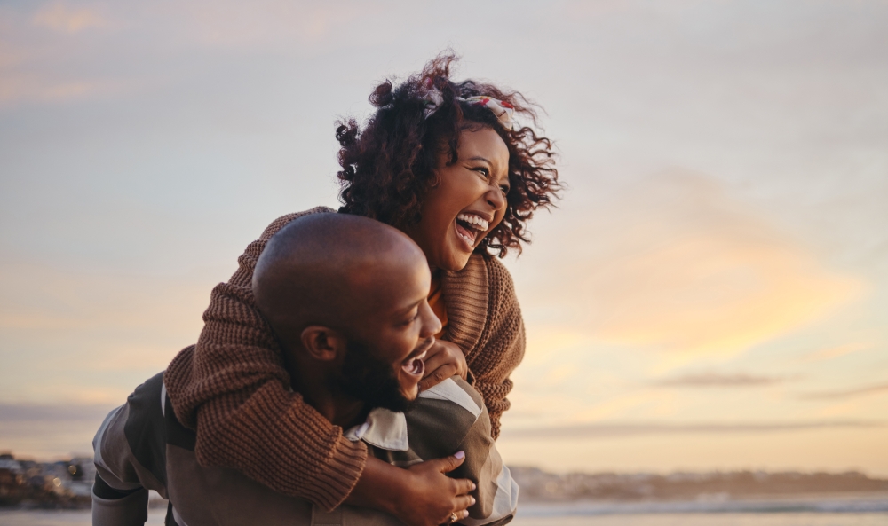 A woman climbs on a man's shoulders, both laughing at sunset. 