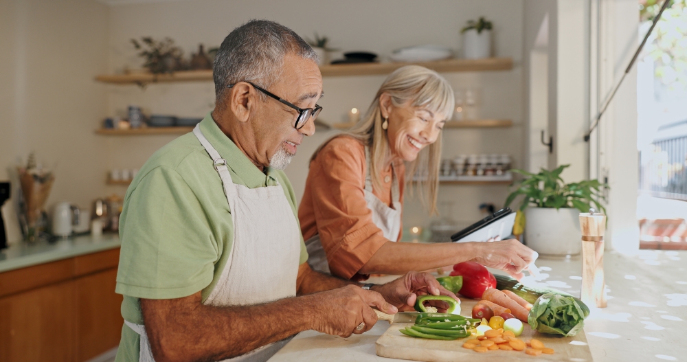 An older couple wearing aprons chop vegetables at a counter in a bright kitchen.
