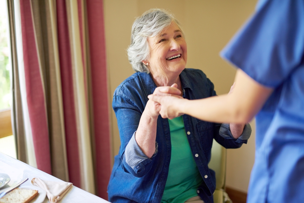 A person off camera holds the hands of an elderly woman as if to help her stand. 
