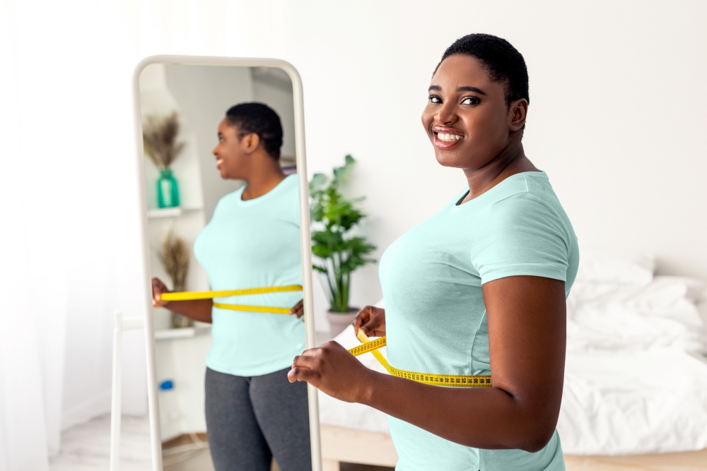 A smiling young woman in a blue tshirt stands in front of a mirror with a yellow tape measurer around her waist.