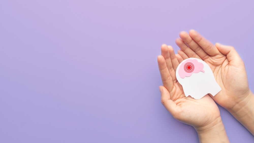 A pair of hands holds a cutout of a head with a red target on the brain, against a light purple backdrop. 
