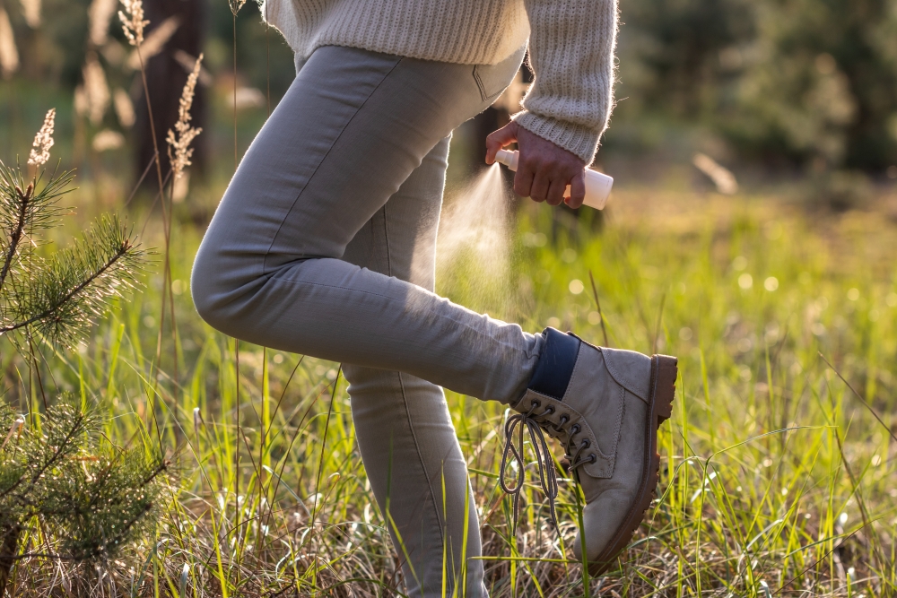 A close-up of a woman in long pants and boots standing in grass and spraying the back of her leg with bug spray. 