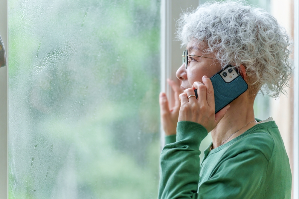 A senior woman holds a phone to her ear, looking out a rain-soaked window.