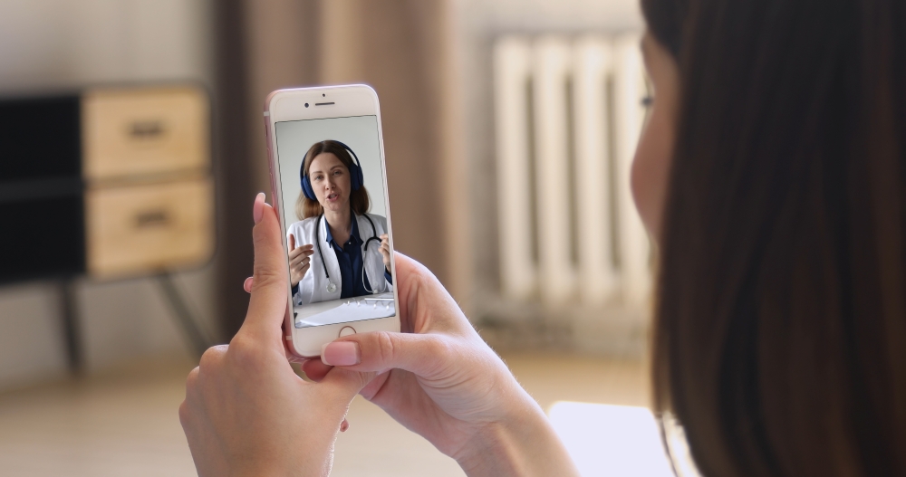 A woman looks at a smartphone screen with a doctor in a labcoat holding a telemedicine appointment.