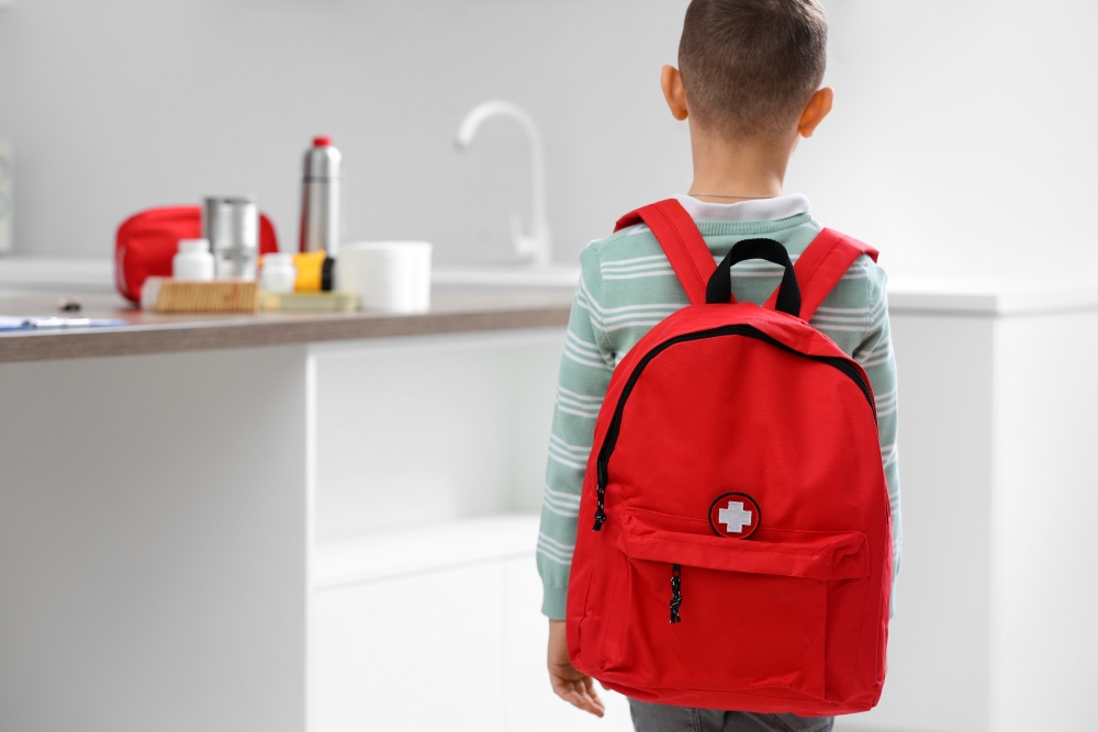 A little boy facing away from the camera wearing a red backpack with a first aid cross on it.