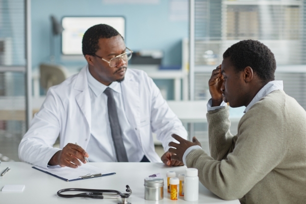 A doctor in a lab coat talks to a patient with his head in his hands. 