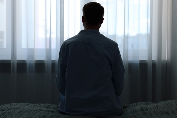 The silhouette of a man in a dark room looking out windows through a sheer curtain. 