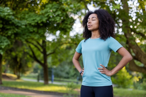 A woman stands outside, taking a deep breath with her hands on her hips. 
