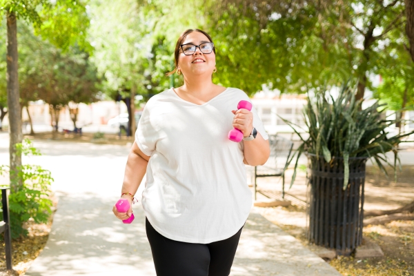 A woman in glasses smiles as she walks outdoors carrying two pink dumbbells.